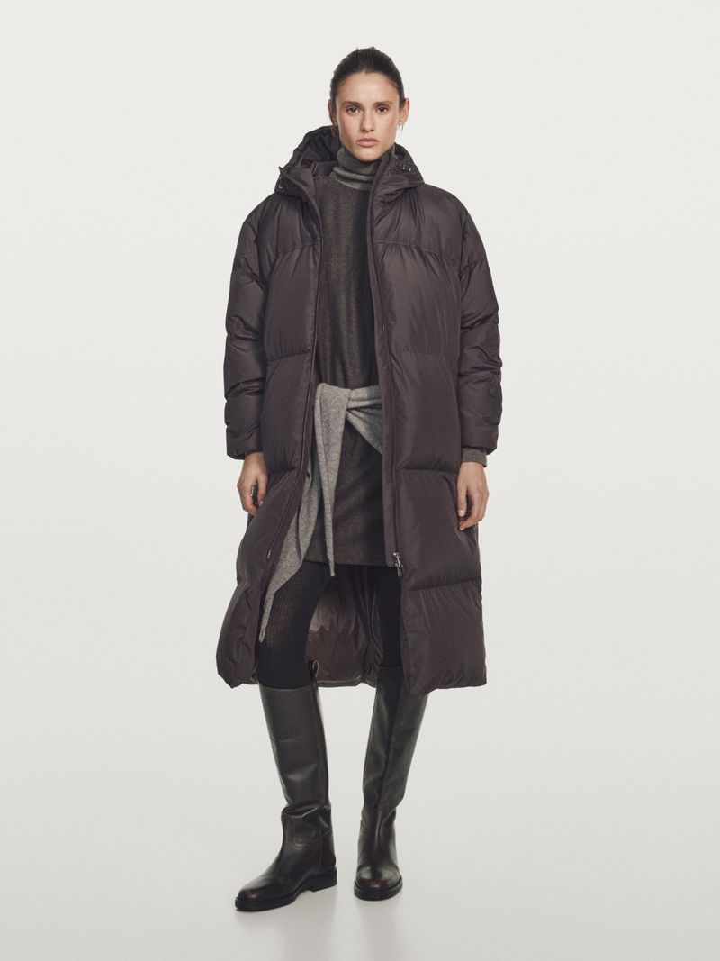 Model Aleyna Fitzgerald wearing a long dark puffer coat, layered knits, and tall black leather boots against a white studio backdrop.