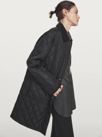Model Aleyna Fitzgerald poses in profile wearing an oversized black quilted coat and dark shirt against a seamless white background.