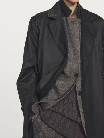 Close-up of a model wearing an open black coat layered over a textured grey wool button-up top and cable knitwear in studio.