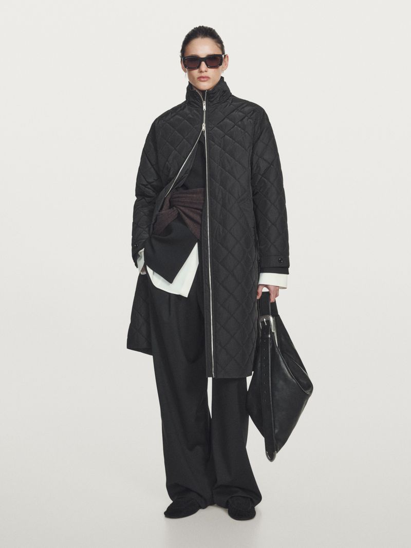 Model Aleyna Fitzgerald wearing a black quilted coat, wide-leg trousers, and sunglasses against a studio backdrop