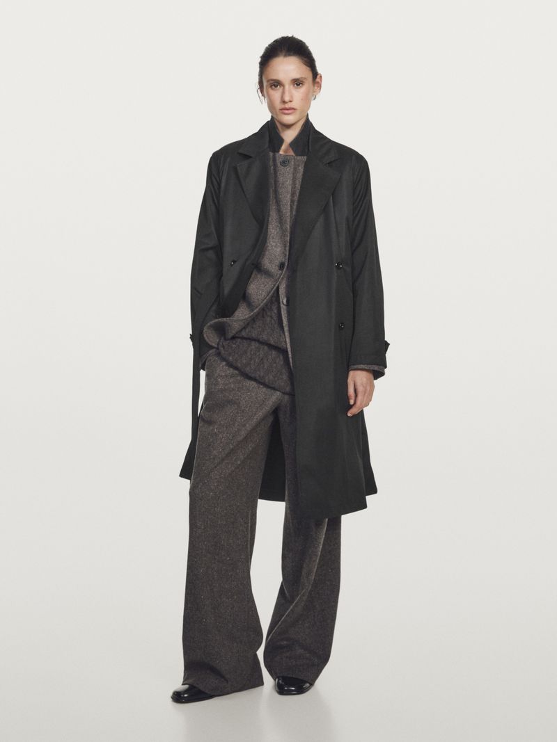 Model Aleyna Fitzgerald wearing an open black coat over a grey textured suit against a light grey studio background.
