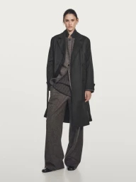 Model Aleyna Fitzgerald wearing an open black coat over a grey textured suit against a light grey studio background.