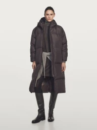 Model Aleyna Fitzgerald wearing a long dark puffer coat, layered knits, and tall black leather boots against a white studio backdrop.
