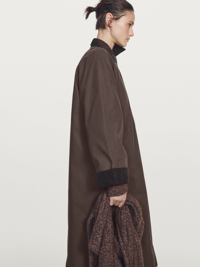 Model Aleyna Fitzgerald wearing an oversized brown coat and holding a knit scarf in profile against a plain white studio background.