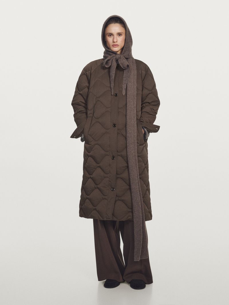 Model wearing a long quilted brown puffer coat with an integrated knit hood scarf and wide-leg trousers against a light backdrop.