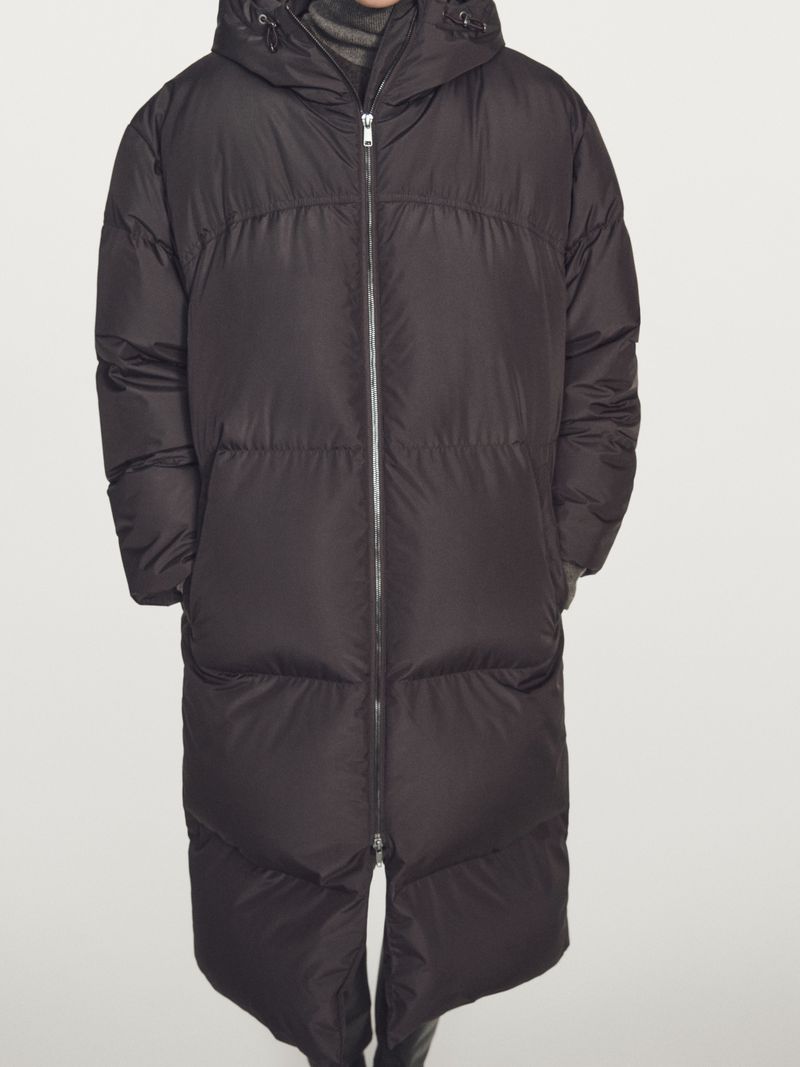 Model wearing a long black hooded puffer coat with a front zipper against a plain seamless studio background.