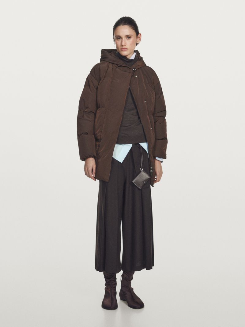 Model Aleyna Fitzgerald poses full-length wearing a brown hooded puffer coat and wide-leg dark trousers against a neutral studio backdrop.
