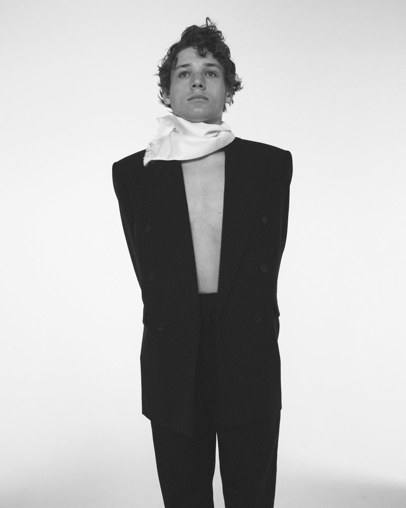 Portrait of a young adult man in a black suit jacket and white scarf, set against a stark white background, striking a confident pose.