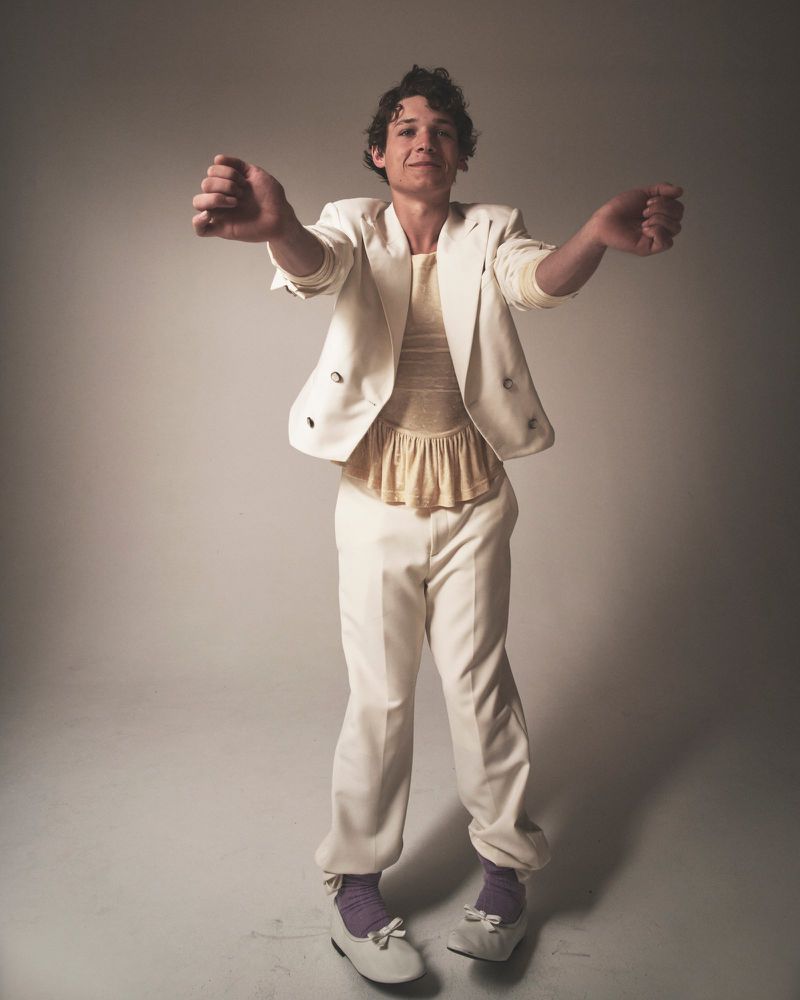 Full-length portrait of a young adult male wearing a white suit with lavender socks and white shoes, posing in a studio setting.