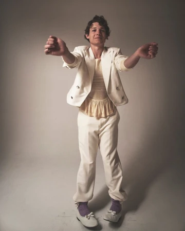 Full-length portrait of a young adult male wearing a white suit with lavender socks and white shoes, posing in a studio setting.