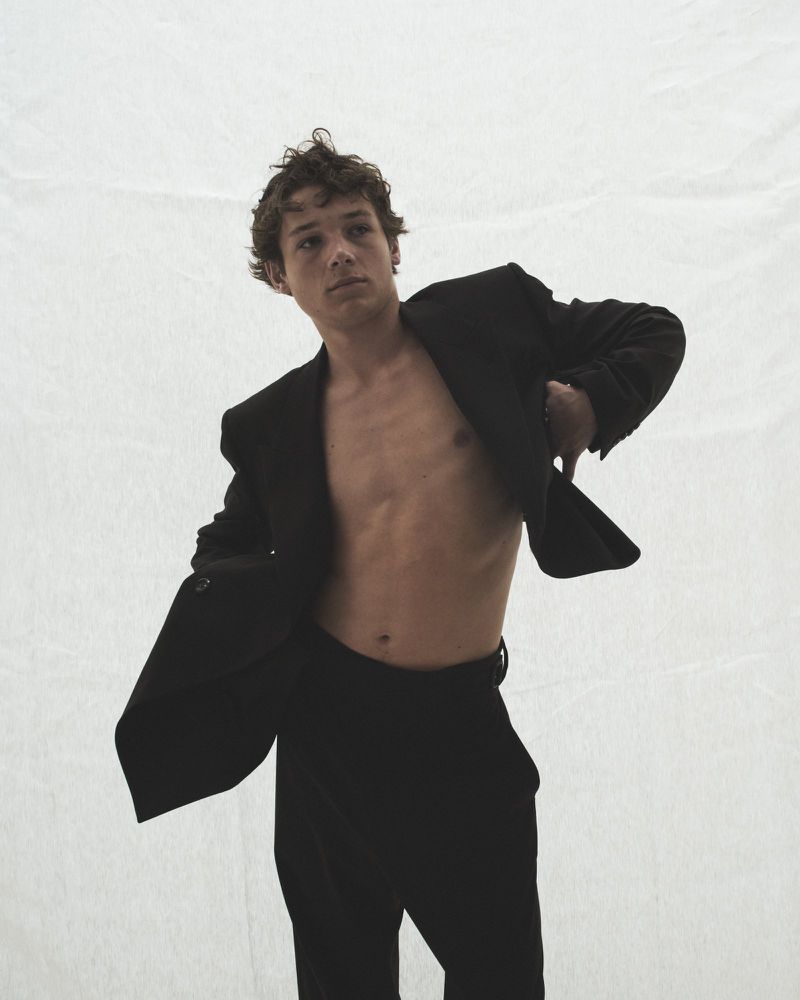 Young adult male model in unbuttoned black suit jacket and pants posing against a white backdrop in a fashion editorial style.