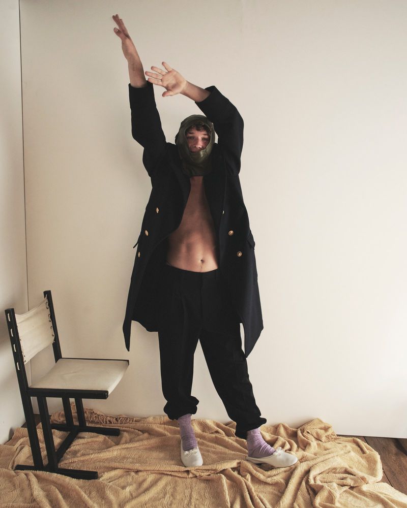 Person in an oversized blazer, dark pants, and slippers poses indoors with raised arms next to a chair, against a neutral background.