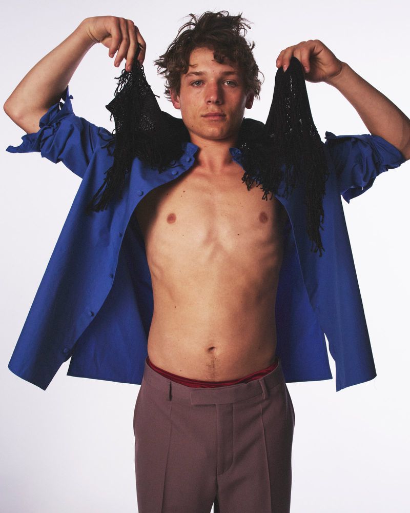 A young adult man poses shirtless in a studio with a blue shirt open, holding black fringed fabrics up with neutral facial expression.