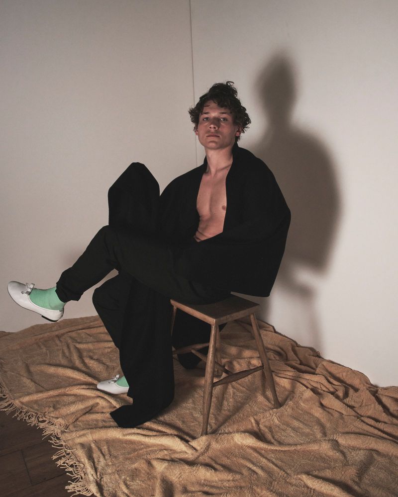 Adult male in casual pose, wearing black robe and green shoes, sitting on wooden stool in a minimalist indoor studio setting.