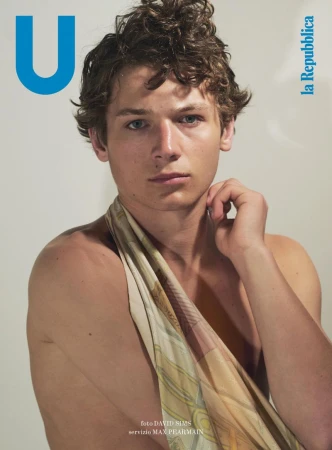 Portrait of a young adult male with curly hair wearing a patterned scarf, posing in front of a plain background, magazine cover