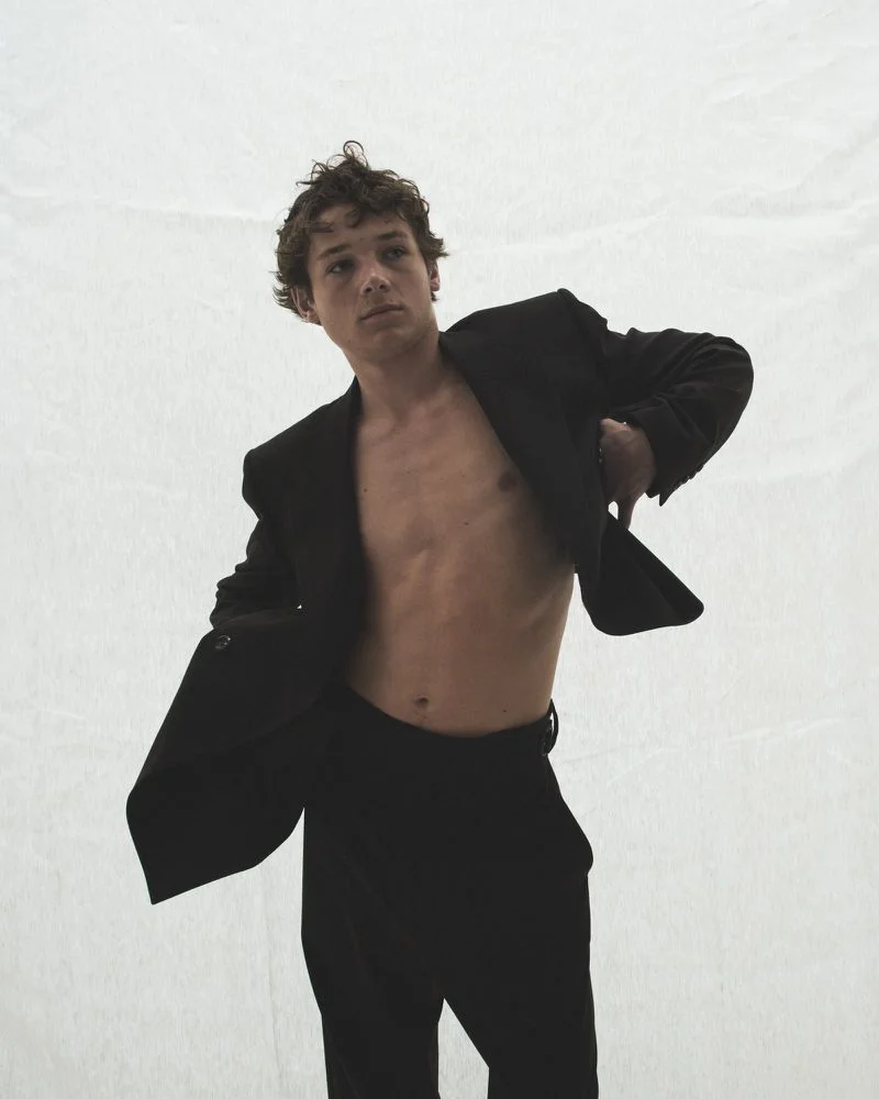 Young man with tousled brown hair wearing an open black suit jacket and dark trousers against a white backdrop.