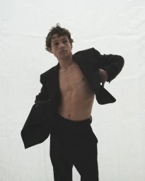 Young man with tousled brown hair wearing an open black suit jacket and dark trousers against a white backdrop.