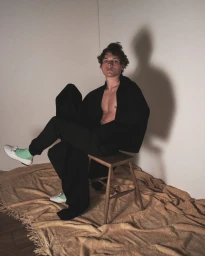 Young man seated on a wooden stool wearing dark trousers and a dark garment, with white shoes and mint green socks.