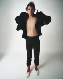 Young man with a bare chest wearing an oversized black jacket, dark trousers, bright red socks, and white shoes against white background