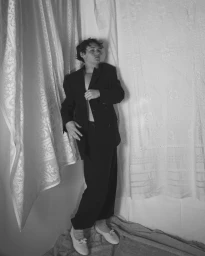 A young man wearing a dark oversized suit standing against a textured draped fabric background.
