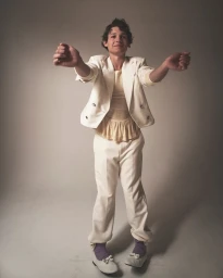A man in an off-white suit, ruffled top, purple socks, and bow-adorned flats posing with arms raised in front of a neutral studio backdrop.