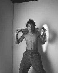 Shirtless young man wearing textured trousers holding a fabric cloth behind his neck in a studio setting