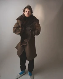 Young man wearing a brown coat with a fur collar and dark trousers standing against a plain grey seamless background.