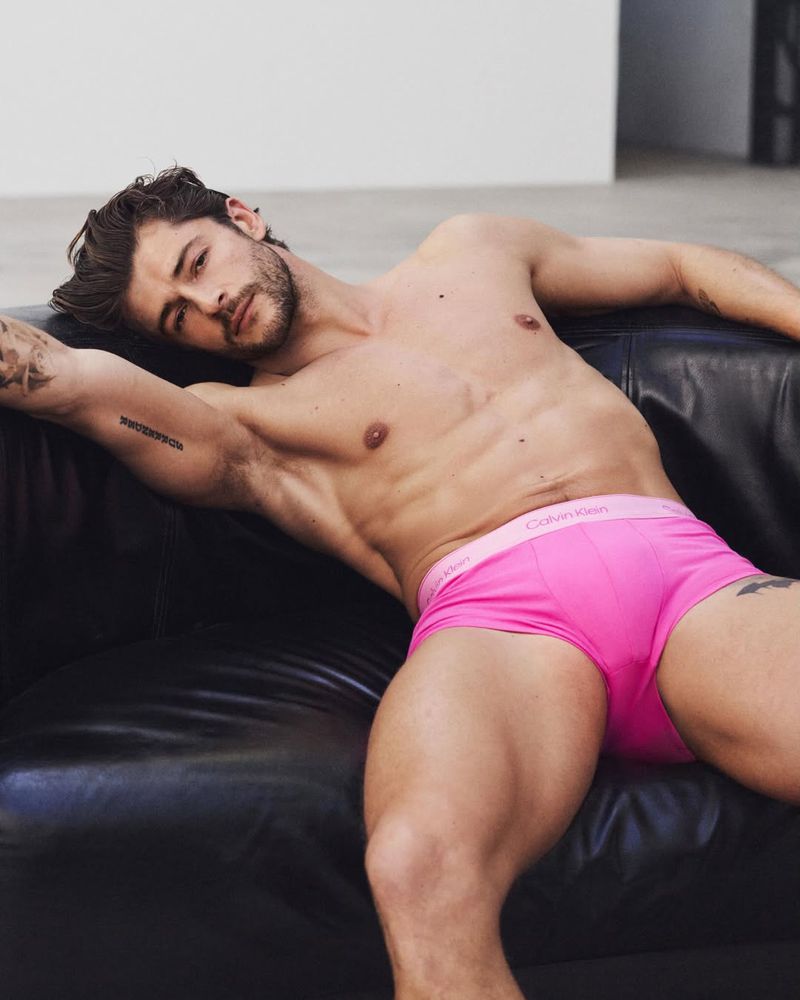 Model Sam Salter reclines on a black leather sofa wearing bright pink Calvin Klein underwear in a studio setting.