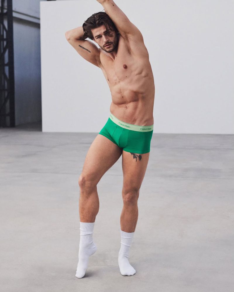 Shirtless male model posing in green Calvin Klein trunks and white crew socks against a neutral studio backdrop.