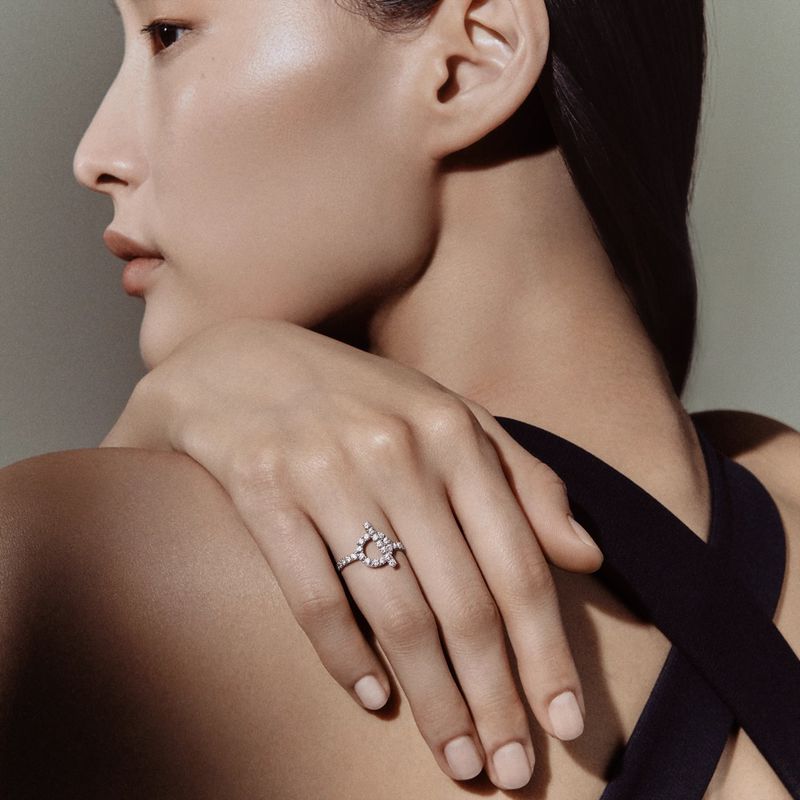 East Asian model in profile resting a hand adorned with a diamond ring on her shoulder against a neutral studio backdrop.