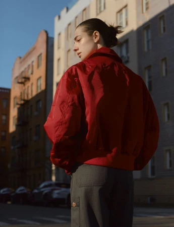 Woman in a red bomber jacket and gray pants stands outdoors in front of urban buildings on a sunny day, captured from the rear.
