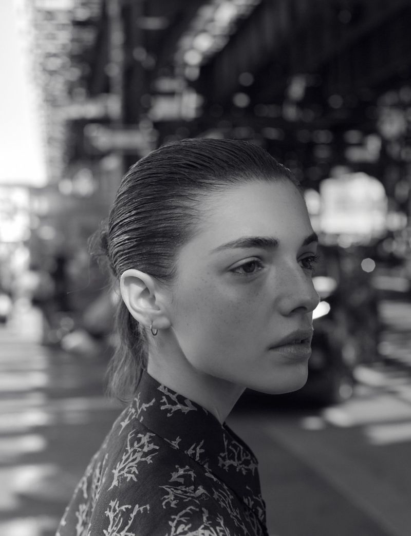 Black and white portrait of a fair-skinned woman in a patterned shirt, photographed outdoors with an urban background in daylight.