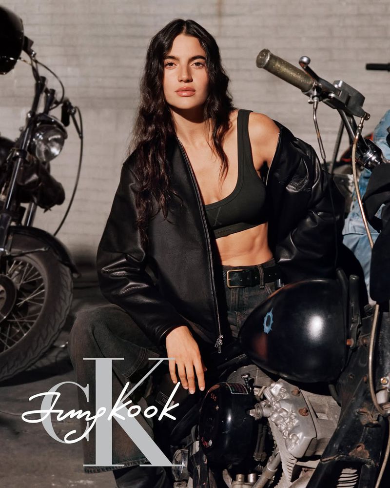 Model in a black leather jacket and crop top seated on a vintage motorcycle with the Calvin Klein Jung Kook logo.