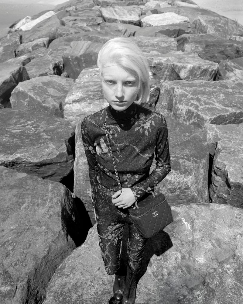 A slender female model in a floral catsuit poses on large rocks at the coast, carrying a Chanel vanity bag in a black and white fashion photograph.