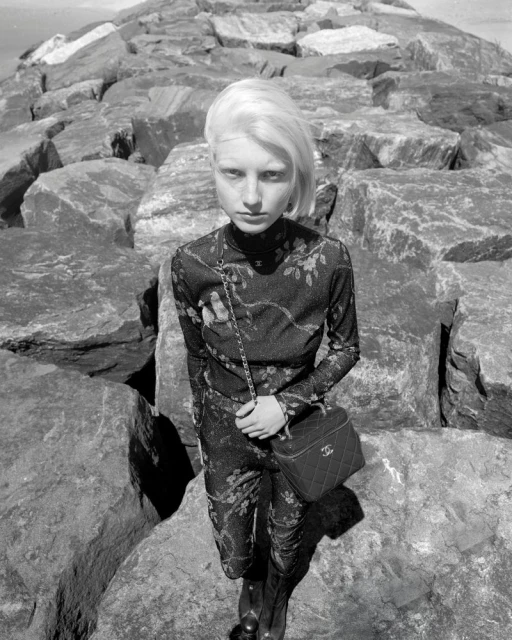 A slender female model in a floral catsuit poses on large rocks at the coast, carrying a Chanel vanity bag in a black and white fashion photograph.