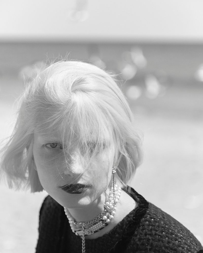 Black and white portrait of an adult woman with blonde hair wearing pearl jewelry and dark lipstick gazing towards the camera.