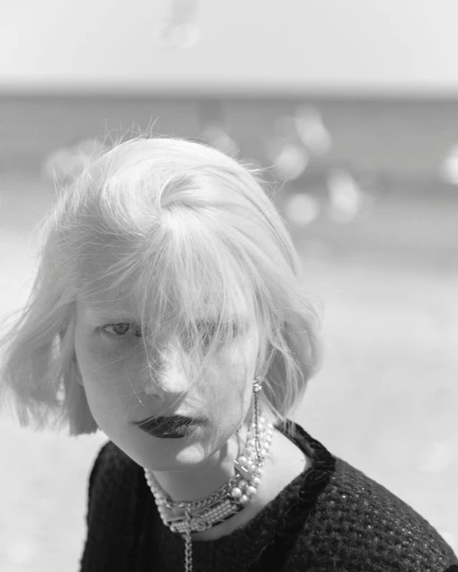 Black and white portrait of an adult woman with blonde hair wearing pearl jewelry and dark lipstick gazing towards the camera.