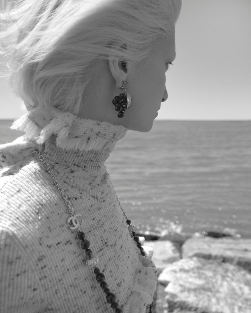 Black and white portrait of a fair-skinned woman with blonde hair in a sweater with a beaded necklace at the beach.
