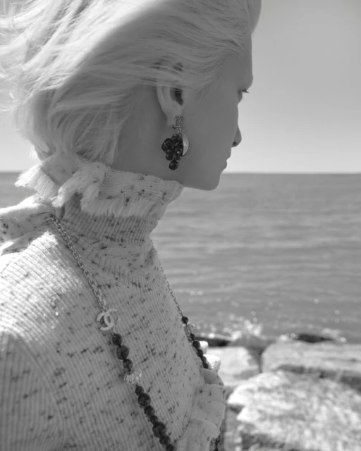 Black and white portrait of a fair-skinned woman with blonde hair in a sweater with a beaded necklace at the beach.