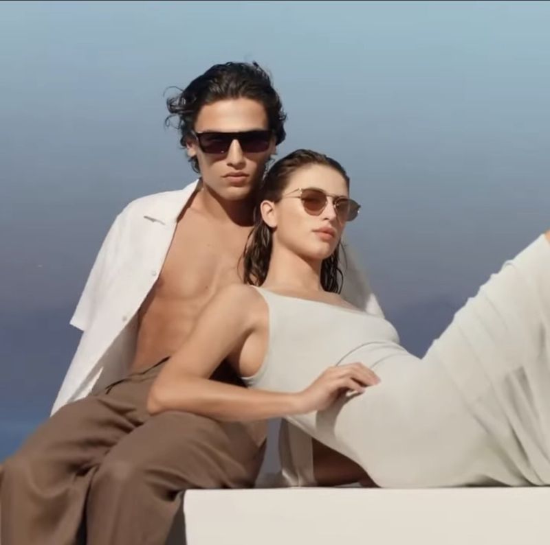 A man and woman wearing sunglasses lounging outdoors in light-colored clothing against a clear blue sky.