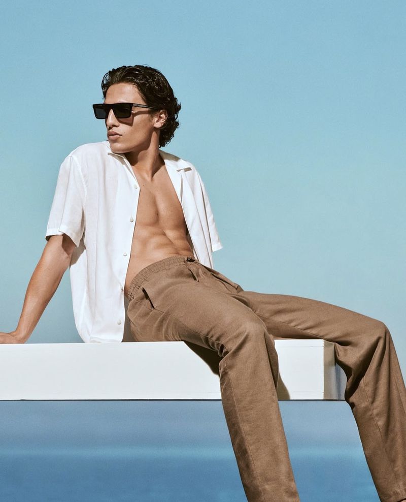 Model wearing dark sunglasses, unbuttoned white shirt, and brown trousers sitting against a clear blue sky background.
