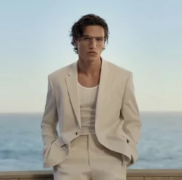Man wearing clear glasses and a cream suit jacket standing outdoors against an ocean backdrop