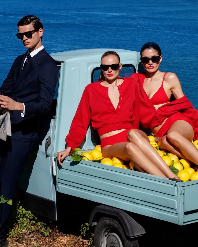 Models in red swimwear and a man in a dark suit pose on a light blue truck bed filled with lemons by the sea.