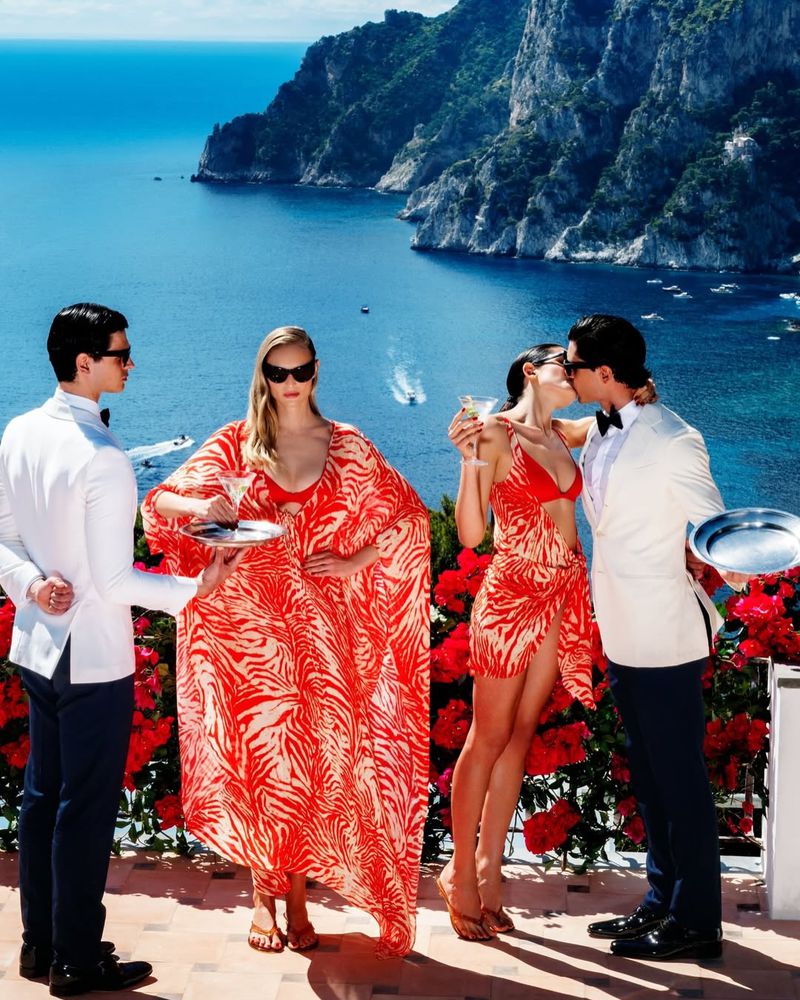 Models in red printed resort wear posed on a cliffside terrace with waiters in white dinner jackets overlooking the sea.