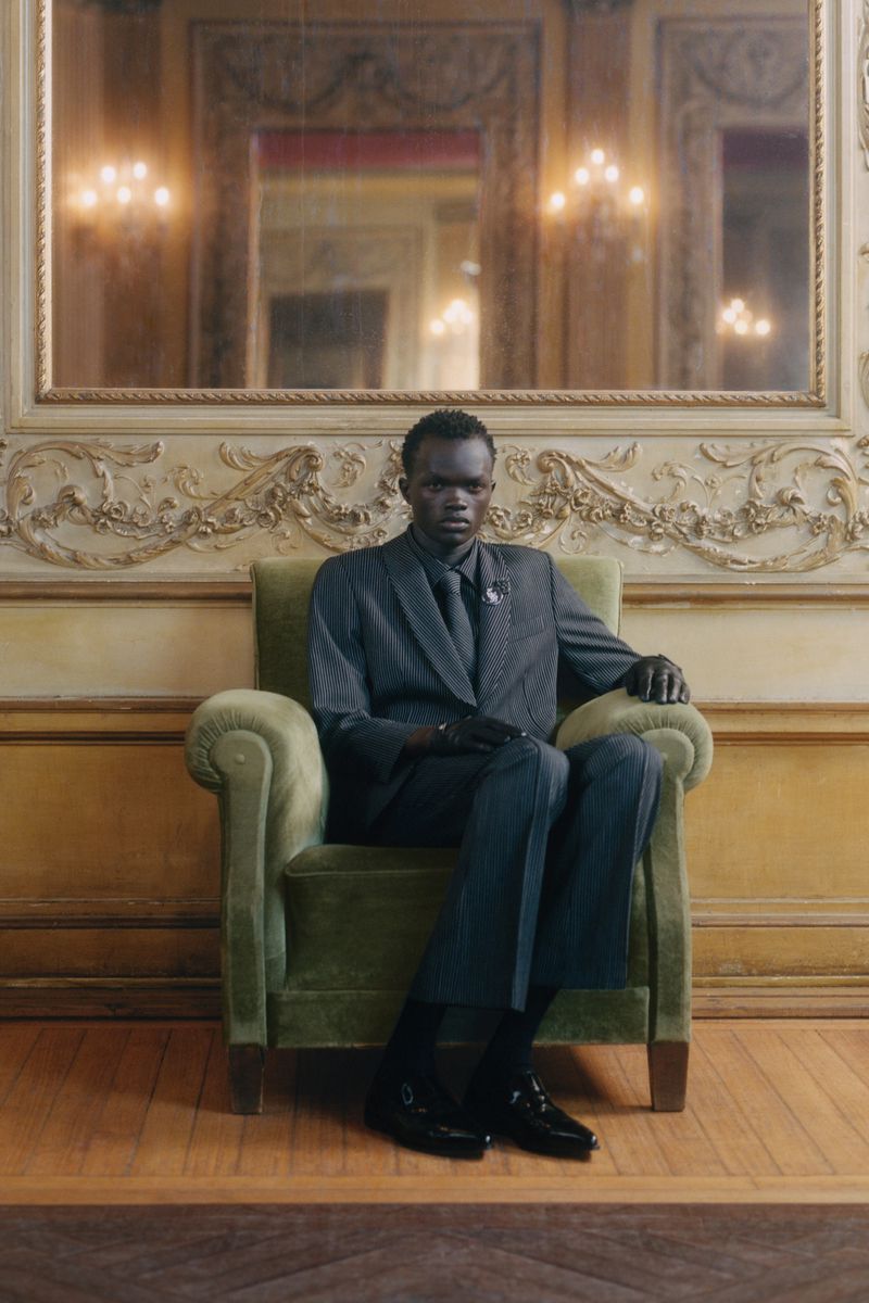 Dark-skinned man in a grey pinstripe suit sits in a green velvet armchair in an ornate room with wood floors and a large mirror.