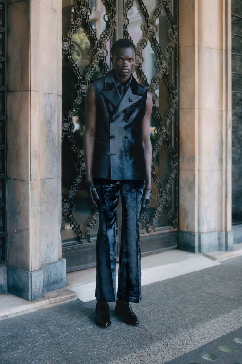 Man poses in sleeveless black suit with velvet pants in front of ornate building doors. Outdoor fashion shot with serious expression.