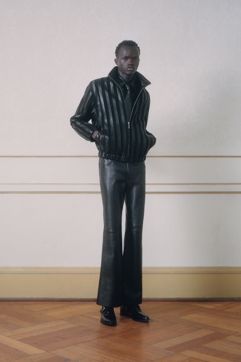 A male fashion model stands in leather jacket and pants in front of a cream colored wall and parquet floor in a fashion forward look.