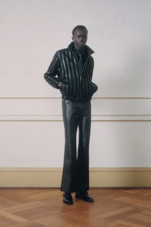 A male fashion model stands in leather jacket and pants in front of a cream colored wall and parquet floor in a fashion forward look.