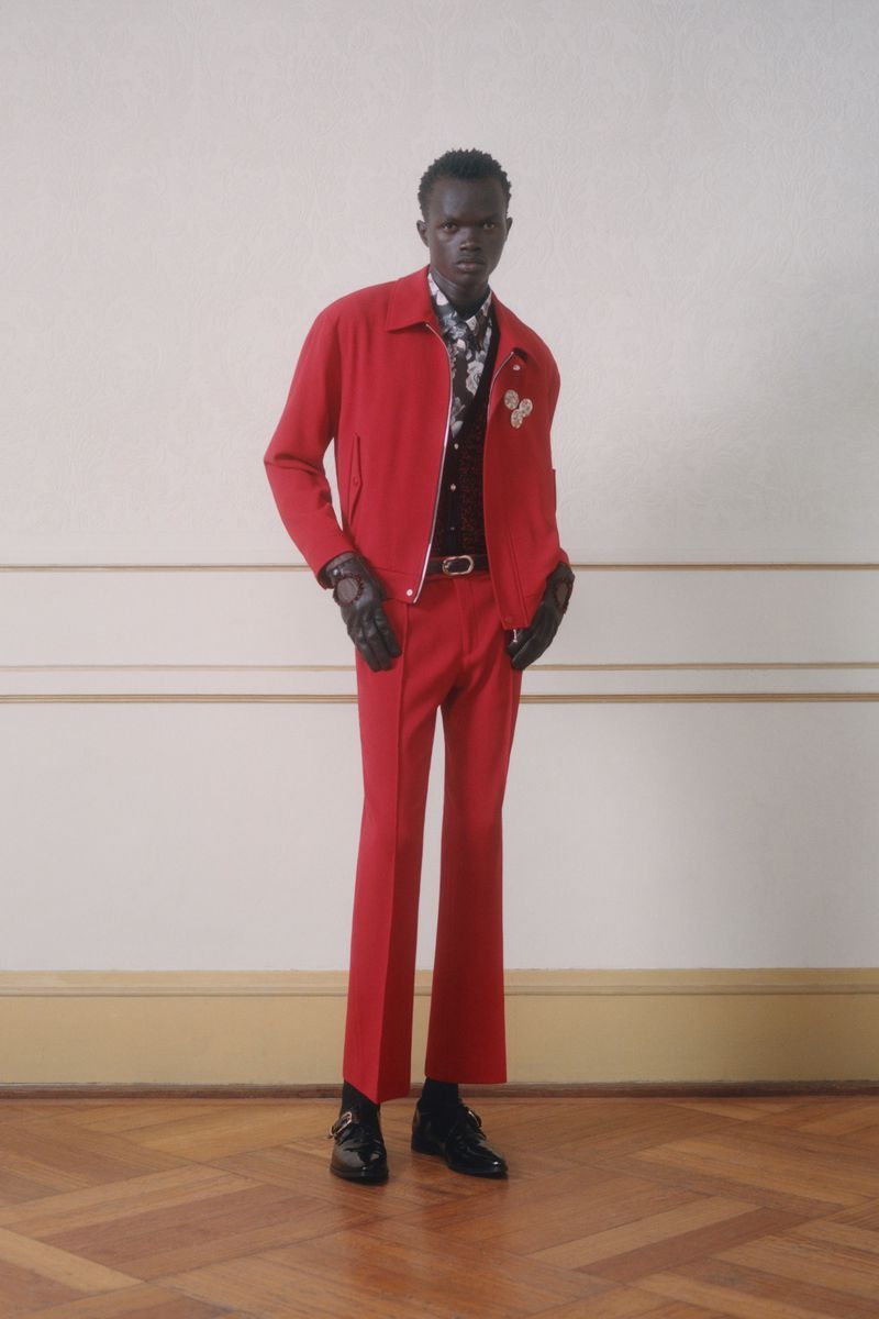 A slender man in a vibrant red jacket and trousers strikes a pose indoors, complemented by black gloves and shoes, blending fashion and elegance.