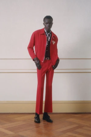 A slender man in a vibrant red jacket and trousers strikes a pose indoors, complemented by black gloves and shoes, blending fashion and elegance.